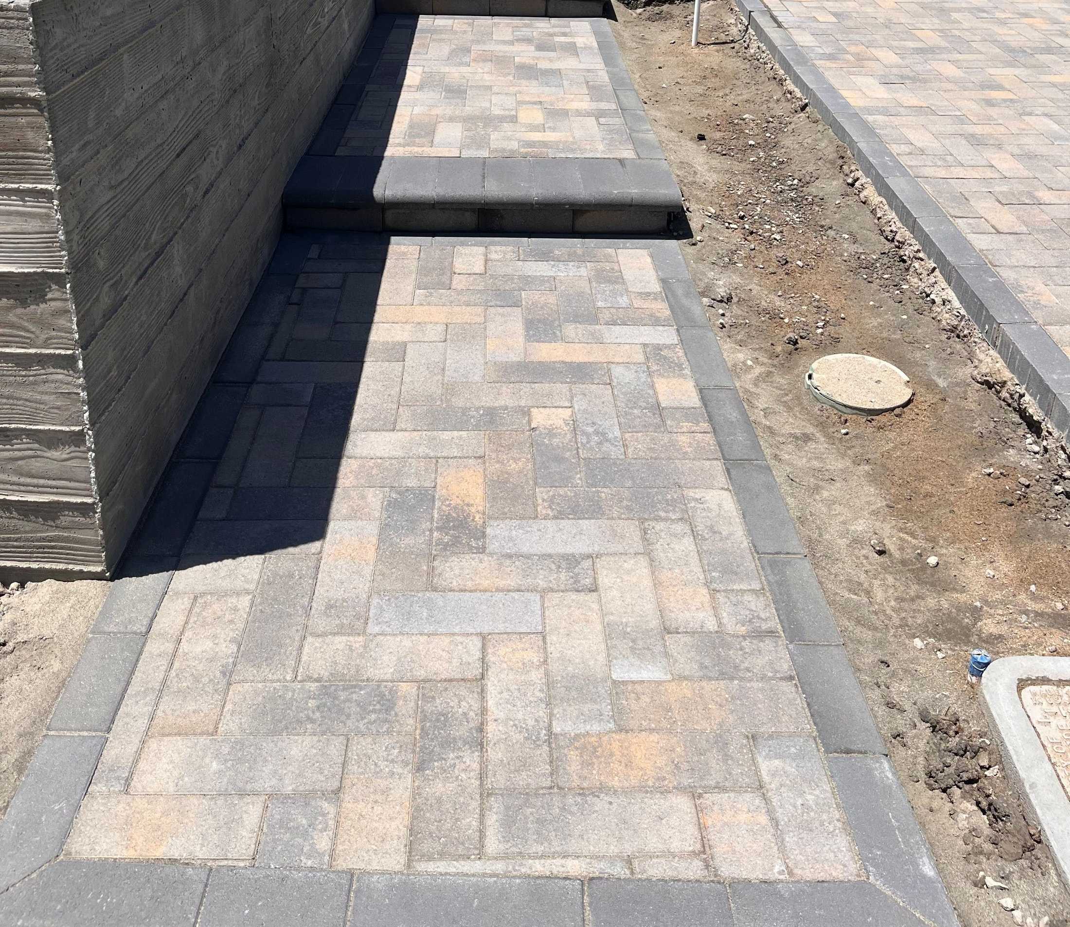 Paver walkway under construction beside a block wall, with exposed soil and unfinished adjoining areas.